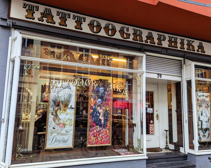 Außenansicht von Tattoographika mit dem ikonischen goldenen Schriftzug und bunten Kunstwerken im Fenster.