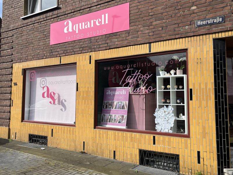 Außenansicht eines Tattoo-Studios mit pinkfarbenem Schild und Schaufensterdekorationen.