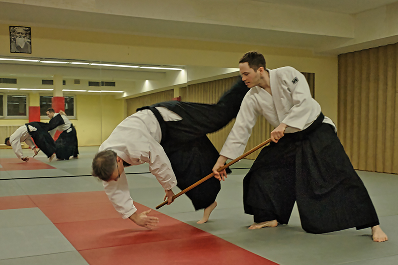Zwei Aikido-Praktizierende üben eine Wurftechnik mit einem Jo (Stab) während des Trainings.