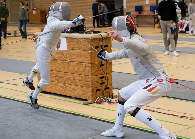 Zwei Fechter im Säbelkampf, einer springt und greift an, während der andere verteidigt, in einer sportlichen Wettkampfumgebung.