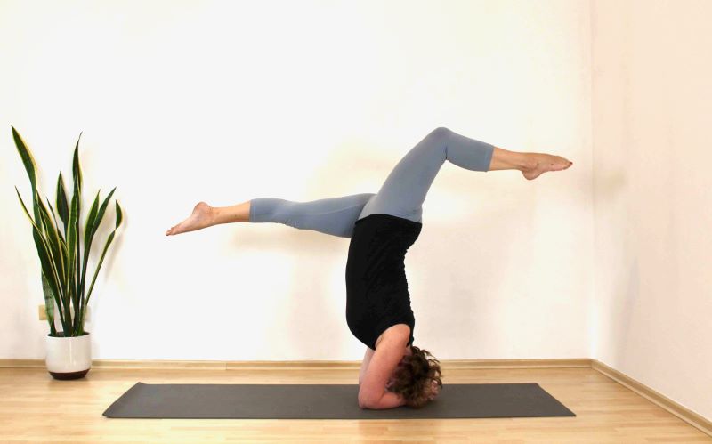 Eine Frau führt im Yoga-Studio einen Kopfstand mit gespreizten Beinen aus, neben ihr steht eine Pflanze.