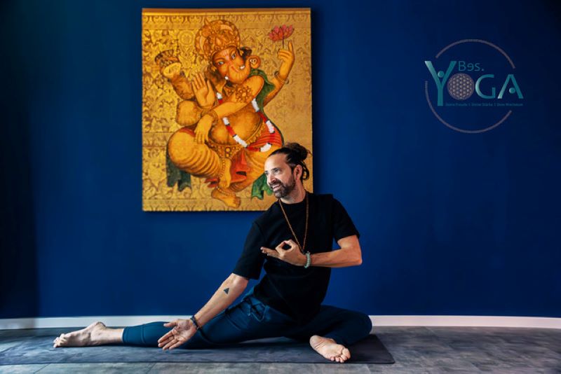 Ein Yogalehrer praktiziert eine sitzende Dehnposition in einem Yoga-Studio vor einem Ganesha-Bild.