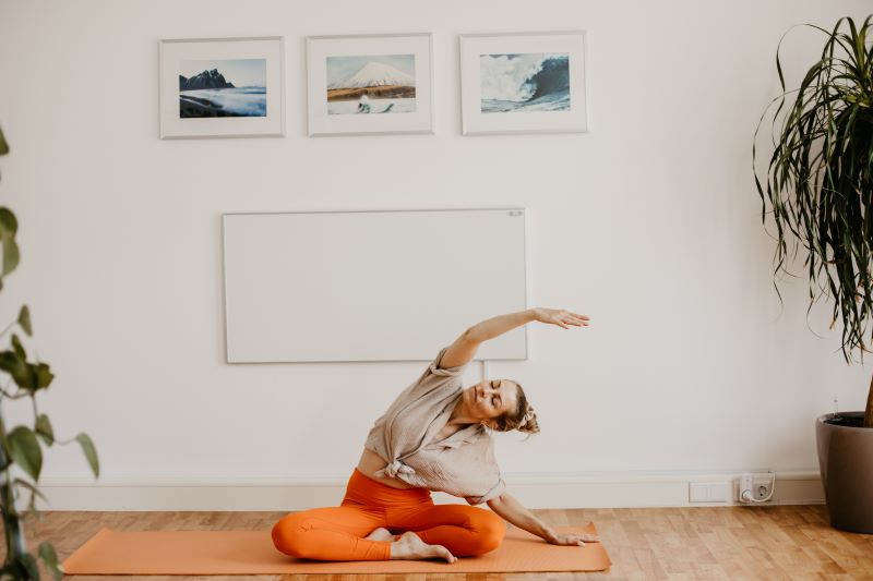 Eine Frau führt eine seitliche Dehnübung auf einer Yogamatte in einem hellen Yoga-Raum aus, umgeben von Pflanzen und Bildern an der Wand.