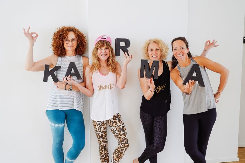 Vier Yogalehrerinnen halten Buchstaben, die zusammen "KARMA" bilden, und lächeln in die Kamera.