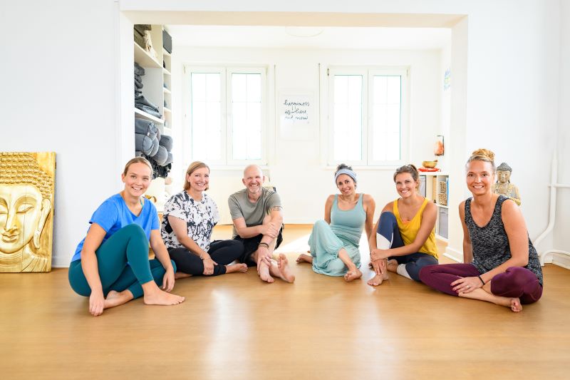 Eine Gruppe von sechs Menschen sitzt entspannt auf dem Boden eines Yoga-Studios und lächelt in die Kamera.