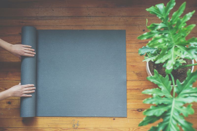 Eine Person rollt eine Yogamatte auf einem Holzboden neben einer grünen Pflanze aus.