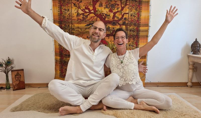 Ein Mann und eine Frau sitzen in weißer Kleidung lächelnd auf einer Yogamatte, die Arme freudig nach oben gestreckt.